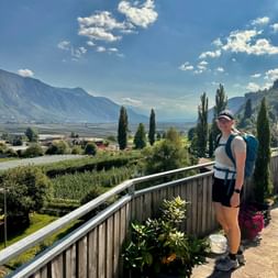 Wanderin mit Rucksack auf Terrasse mit Blick auf Tal zwischen Meran und Gardasee. Zypressen und Berge unter blauem Himmel sichtbar.
