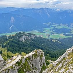 Wanderer auf einem felsigen Bergpfad am Wendelstein mit Panoramablick auf bewaldete Gipfel, grüne Täler und ein bayerisches Dorf.