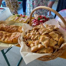 Buffettisch mit Weidenkorb voller Gebäck, Baguettes, Käseplatte und Erdbeerdessert. Menschen in Sommerkleidern stehen im Hintergrund.