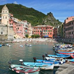 Hafen von Vernazza mit bunten Fischerbooten, pastellfarbenen Häusern, Kirchturm und grünen Hügeln. Strand mit Badenden und Booten im türkisfarbenen Wasser.
