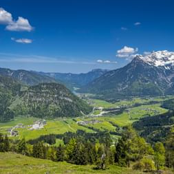 Panoramic view from Buchensteinwand in the Kitzbühel Alps showing green valley with villages, forested mountains, and snow-capped peaks under blue sky.