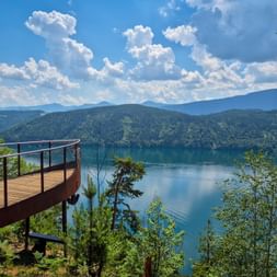 Geschwungene Aussichtsplattform mit Blick auf den Millstätter See mit türkisfarbenem Wasser, bewaldeten Bergen und weißen Wolken.