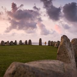 Ales Stenar Megalithdenkmal mit stehenden Steinen in Schiffsformation auf grünem Gras unter dramatischem Wolkenhimmel bei Sonnenuntergang.