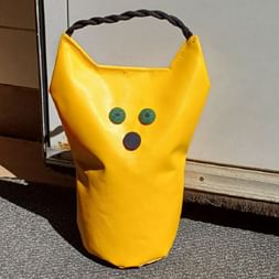 Yellow fabric door stopper with black braided handle, featuring two green circular eyes and a brown circular mouth, positioned on concrete.