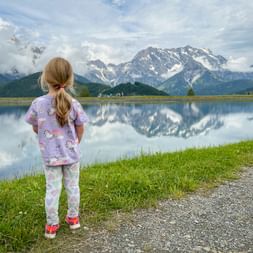 Child with blonde ponytail standing by Prinzensee lake in Pinzgau, viewing snow-capped mountains reflected in calm water.
