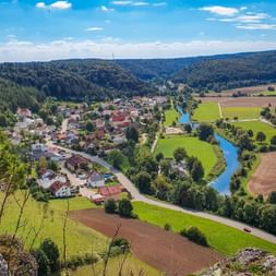 Luftaufnahme des Dorfes Arnsberg im Altmühltal mit blauem Fluss, der sich durch grüne Wiesen und Felder schlängelt, umgeben von bewaldeten Hügeln.