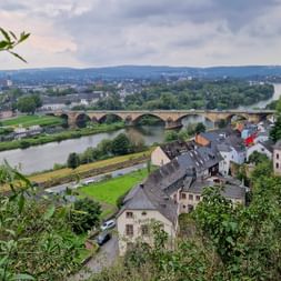 Erhöhte Ansicht von Trier mit historischer Steinbrücke über die Mosel, Wohngebäuden im Vordergrund und grünen Hügeln im Hintergrund.