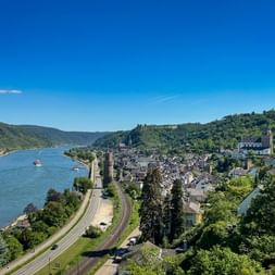 Luftaufnahme von Oberwesel am Rhein mit mittelalterlichen Türmen, Weinbergen an den Hängen und Schiffen auf dem Wasser unter blauem Himmel.