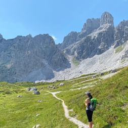 Wanderer mit grünem Rucksack auf Bergweg im Salzkammergut. Grüne Almwiese führt zu den markanten Bischofsmützen unter blauem Himmel.