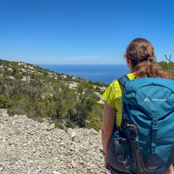 Female hiker with blue Vaude backpack viewing the Adriatic Sea coastline in Kvarner Bay, Croatia. Rocky hillside with vegetation and blue sky.