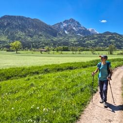 Wanderin mit Rucksack und Stöcken auf Feldweg durch grüne Wiese bei Piding, mit bewaldeten Bergen unter blauem Himmel.