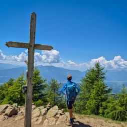 Wanderin mit blauem Rucksack steht am hölzernen Gipfelkreuz der Millstätter Alpe und blickt auf Bergketten unter blauem Himmel mit weißen Wolken.