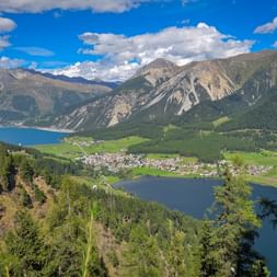Aerial view of Reschensee and Haidersee lakes surrounded by mountains. A village nestles between the lakes with green meadows and forests.