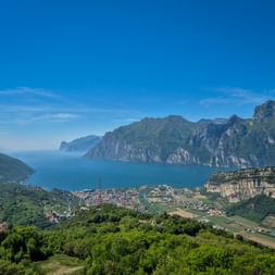 Aerial view of Lake Garda surrounded by mountains, with towns and green hillsides in the foreground under a clear blue sky.