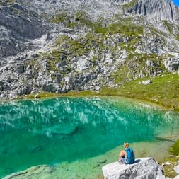 Hiker with blue backpack sitting on rock by turquoise mountain lake in Allgäu-Lechtal Alps, surrounded by dramatic rocky cliffs and green slopes.