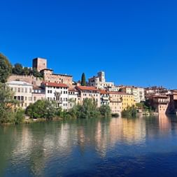 Historische Stadt Bassano del Grappa mit bunten Häusern am Fluss, mittelalterlichem Turm und überdachter Holzbrücke unter blauem Himmel.