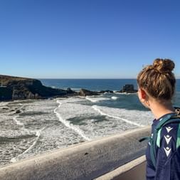 Wanderin mit Rucksack betrachtet felsige Küste bei Zambujeira do Mar, Alentejo. Wellen brechen an dunklen Klippen unter blauem Himmel.