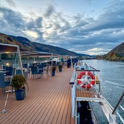 Deck of a river cruise ship on the Danube with dining tables and chairs. Mountains with autumn foliage line both sides under a cloudy sky.