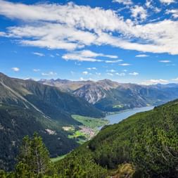 Panoramic view of Reschensee lake nestled in an Alpine valley, surrounded by forested mountain slopes and distant peaks under a blue sky.