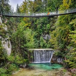 Suspension bridge labeled Kranjska Gora spanning a forested gorge above a waterfall flowing into a turquoise pool surrounded by green vegetation.