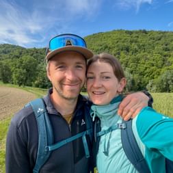 Two hikers taking a selfie in Piedmont with green hills and fields. Man with cap and woman in turquoise jacket, both wearing backpacks.