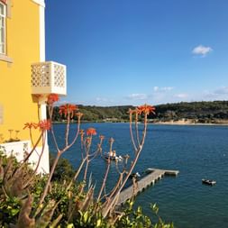 Gelbes Gebäude mit rot blühenden Aloen mit Blick auf einen blauen Fluss mit Holzsteg und Booten in Vila Nova de Milfontes, Portugal.