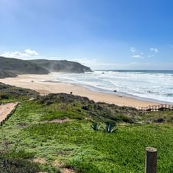 Praia do Amado Strand mit goldenem Sand, rollenden Wellen und Küstenklippen. Grüne Vegetation und Holzstege im Vordergrund.