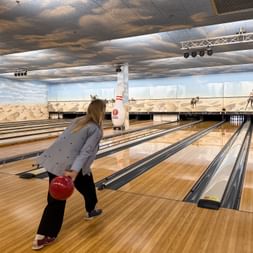 Frau namens Chrissi beim Bowlen in einer Bahn mit Wüsten-Wandbild im Hintergrund. Sie hält eine rote Kugel und bereitet sich auf den Wurf vor.