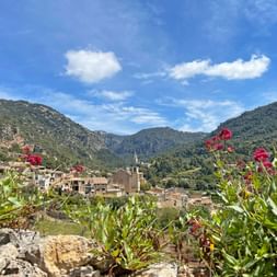 View of Valldemossa village with stone buildings and church tower, framed by pink flowers and green plants, mountains in the background.