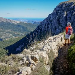 Two hikers on stone-walled mountain path overlooking Serra de Tramuntana valley with sea view. Rocky cliffs and Mediterranean vegetation visible.