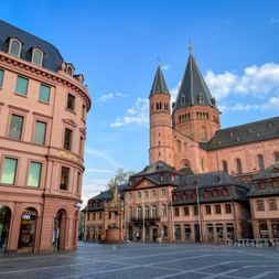 Mainzer Dom mit markanten roten Sandsteintürmen erhebt sich über einem historischen Marktplatz mit traditionellen Gebäuden unter blauem Himmel.