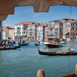 Venice Grand Canal with gondolas and water taxis navigating between historic palazzos. Colorful buildings line the waterway under blue sky.