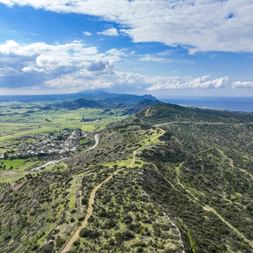 Luftaufnahme der Region Famagusta auf Zypern mit grünen Hügeln, gewundenen Wegen, landwirtschaftlichen Ebenen und fernen Bergen unter bewölktem Himmel.