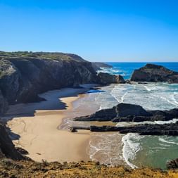 Sandstrand Praia dos Alteirinhos bei Zambujeira do Mar mit dunklen Felsformationen, türkisfarbenen Wellen und steilen Klippen unter blauem Himmel.