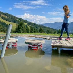 Child on wooden platform at Montelino's water play area in Saalbach-Hinterglemm with mountain views and stepping stones in shallow water.