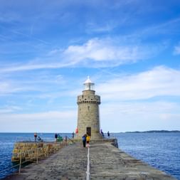 Steinleuchtturm am Ende eines Kopfsteinpflasterpiers in St. Peter Port. Besucher gehen auf dem Pier mit blauem Meer und fernen Inseln.