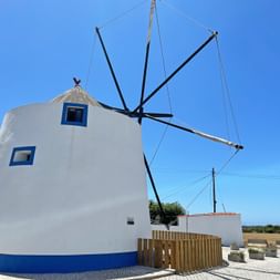 Weiße zylindrische Windmühle mit blauem Rand und Fenstern in Longueira. Schwarze Holzflügel ragen von der Struktur gegen einen klaren blauen Himmel.