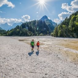 Two hikers walking on a dry lakebed at Lake Jasna near Kranjska Gora, with forested mountains and blue sky in the background.
