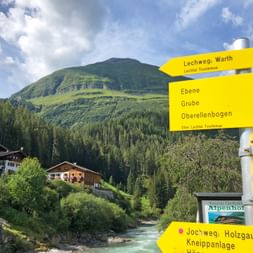Gelbe Wanderwegweiser zeigen Lechweg-Richtungen nach Warth, Holzgau und anderen Zielen, mit Alpendorf, Fluss und grünen Bergen.
