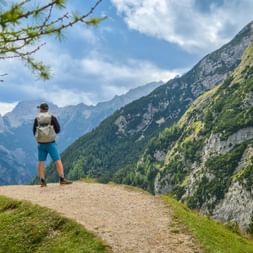 Hiker with backpack standing on mountain path at Vršič Pass in Julian Alps, overlooking Soča Valley with dramatic peaks and forested slopes.