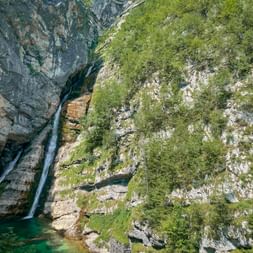 Aerial view of Savica Waterfall cascading down rocky cliffs into a turquoise pool, surrounded by lush green vegetation in the Julian Alps.