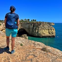 Hiker with backpack standing on rocky cliff edge overlooking turquoise sea and natural rock arch along the Algarve coast in Portugal.