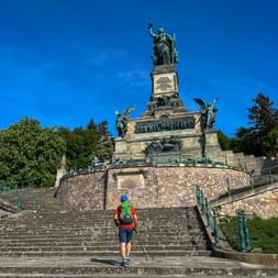 Wanderer mit rotem Rucksack steigt Steintreppen zum Niederwalddenkmal hinauf, einem hohen Denkmal mit Bronzestatuen vor blauem Himmel.