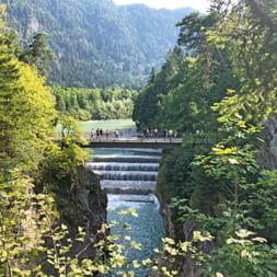 Lechfall waterfall near Füssen with cascading water, viewing bridge with visitors, framed by green trees and forested mountains in background.