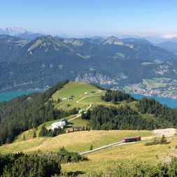Panoramic view from Schafberg mountain showing green alpine meadows, winding paths, turquoise lake, and mountain ranges in Salzkammergut.