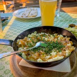 Black pan with Käsespätzle topped with crispy onions and herbs on a wooden board. Beer glass and salad plates visible on a green tablecloth.