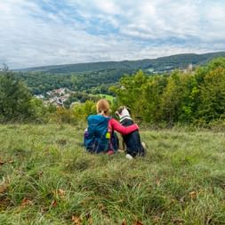 Two hikers with backpacks and a dog sitting on a grassy hillside near Pappenheim, overlooking the Altmühltal valley with forested hills.