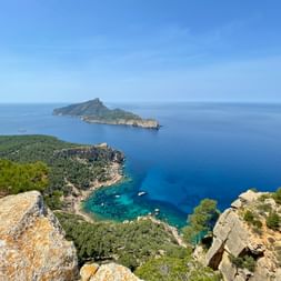 View from rocky cliffs near Sant Elm on Mallorca's GR221 trail, showing turquoise bay, green peninsula, and island in blue Mediterranean Sea.