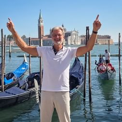 Walter Mirz with raised arms celebrating at Venice waterfront with gondolas and San Giorgio Maggiore church in background.