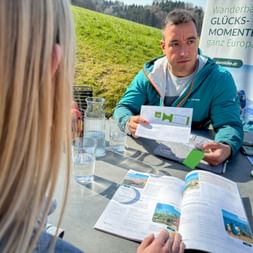 Eurohike-Berater in türkisfarbener Jacke präsentiert Reisebroschüren einem Kunden an einem Tisch im Freien mit Wassergläsern und grüner Wiese.